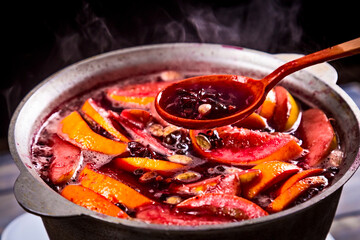 Big kettle pot of traditional hot steaming Christmas drink gluhwein mulled red spiced wine boiling with apples and citrus with wooden spoon on black background. Christmas party.