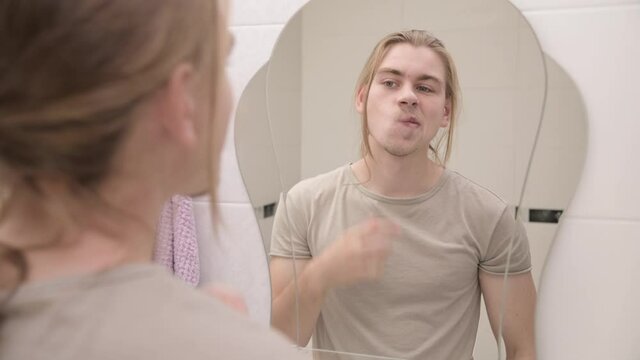 A young man with long hair stands in front of a mirror in the bathroom and examines his face. Self-care and self-care for men