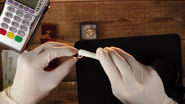 Owner of pawnshop cleaning gold ring with a ruby by a brush. Close-up of goldsmith's work place with jewellery and payment terminal on transparent glass table. Concept of buying up of precious metals.