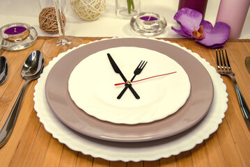 Table setting. Beautiful dishes on a wooden napkin with a crystal glass, candles and flowers. Hands of the clock - lunch time. Close up.