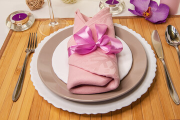 Table setting. Beautiful dishes and a pink napkin with a satin ribbon on a wooden stand with a crystal glass, candles and flowers. White background.