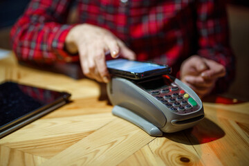 Fototapeta premium Contactless payment. Nfs payment by phone. Client of cafe or restaurant holding smartphone over electronic payment machine while using mobile app to pay. Non cash payment. NFC technology