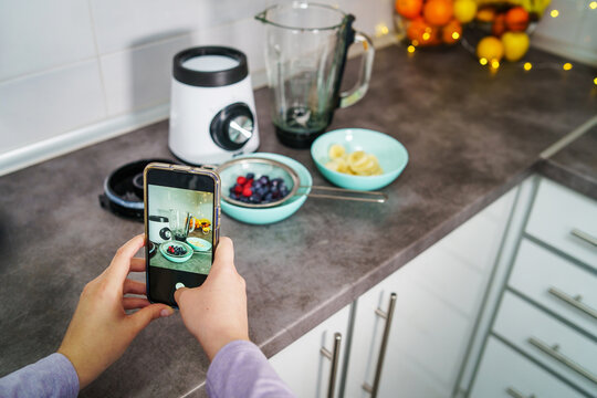 Hands of unknown female woman taking photo with mobile phone banana in the kitchen with raspberry and blueberries in a plate making organic fruit smoothie - healthy eating concept