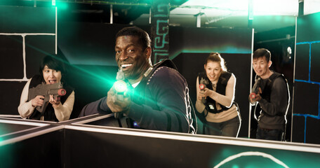 Fototapeta na wymiar Emotional African man with laser pistol playing laser tag with friends on a dark labyrinth