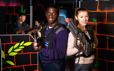 Happy glad positive African man and Caucasian woman standing back to back with laser pistols in dark lasertag room.