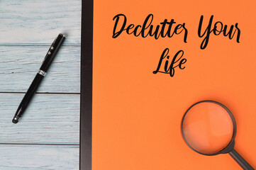 Top view of magnifying glass,pen and orange paper writing with text ' Declutter Your Life ' on wooden background.