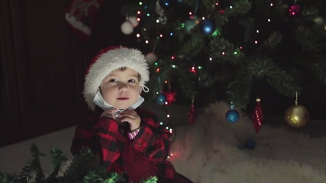 coronavirus, covid-19 in the new year. a little girl in a red Santa Hat and mask is sitting under the Christmas tree at home.