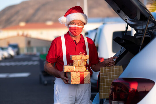 A Senior Man Shopping For Christmas In Santa Hat And Suspenders, Wearing Medical Mask To Avoid Covid Coronavirus Infection, Holding A Shopping Bag Full Of Gifts While Standing In The Mall Parking Lot