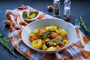 Stew, braised pork ribs with potatoes and carrots in a gray bowl on a black concrete background.
