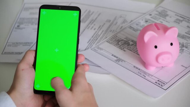 Phone with green screen and piggy Bank payment receipts, tax documents on white background top View . smartphone chromakey swipes in different directions. copy space. High quality 4k footage
