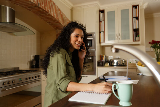 Mixed race woman talking on phone and writing in kitchen