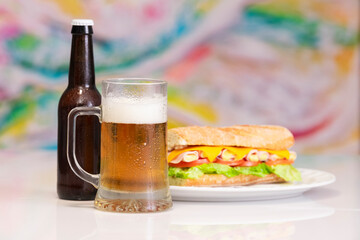 view of a cold mug of beer with out of focus beer bottle and an appetizing sub sandwich