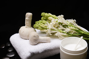 Spa composition with bunch of tuberose flowers and herbal, ball on towel, with spoon cream in bowl pile of black stones