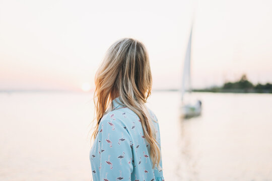 Beautiful blonde young woman in blue dress on pier on background of sailboat on the sunset