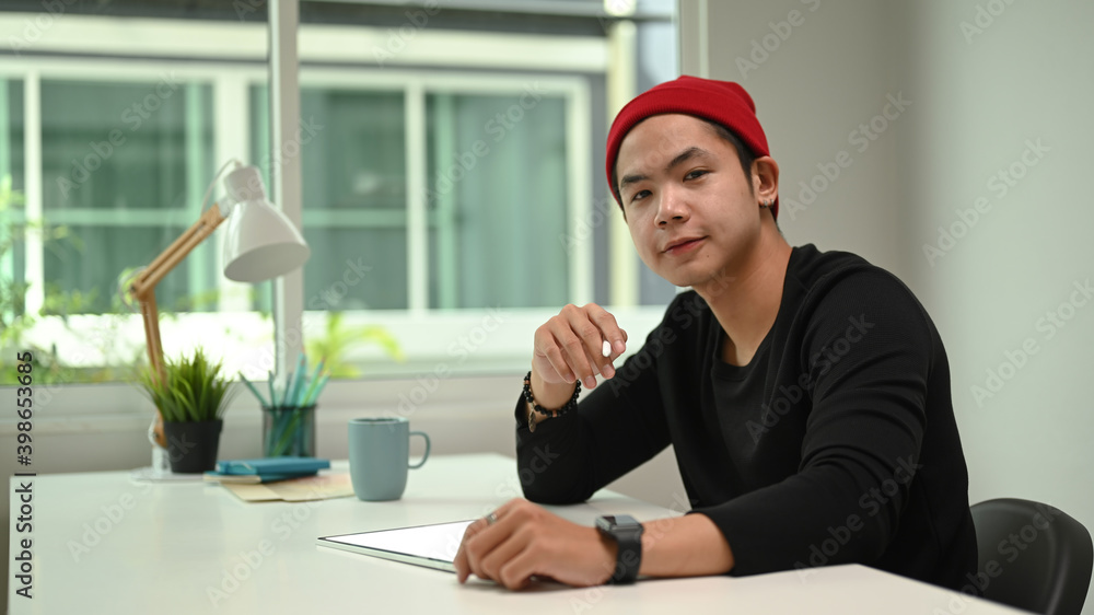 Poster graphic designer in red wool hat working on digital tablet and looking at camera while sitting at hi