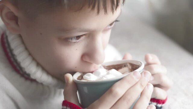 A boy in a warm sweater drinks hot cocoa with marshmallows.