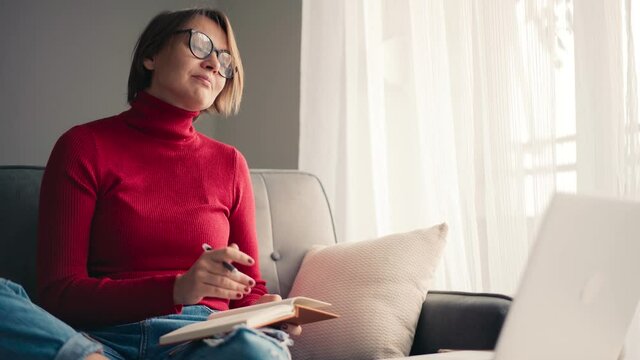 A middle-aged woman in glasses talking on video call on the laptop and making notes in her notebook. Online consultation with a psychologist. Distance learning.