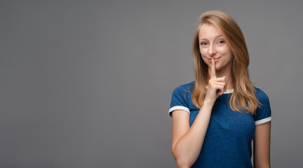 Pretty young blonde woman keeps finger on lips, making hush gesture on gray background. Shh, silence concept