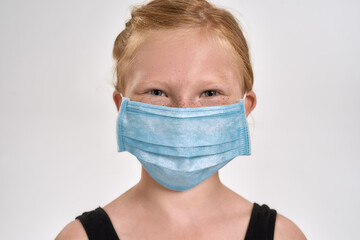 Portrait of pretty little red haired sportive girl in sportswear wearing medical mask, smiling at camera while standing isolated over white background