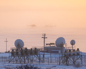 Industrial arctic landscape. Satellite dishes. In the distance, a helicopter is landing on an airstrip in the tundra in the Arctic. Polar aviation. Cold frosty and windy winter weather. Bad visibility