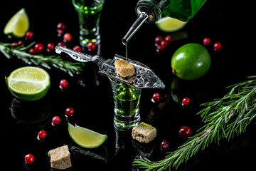 Obraz na płótnie Canvas absinthe shots with sugar cubes. absinthe poured into a glass. bottle of absinthe with brown sugar, cranberries and lime, stainless steel spoon isolated on black background. space for text