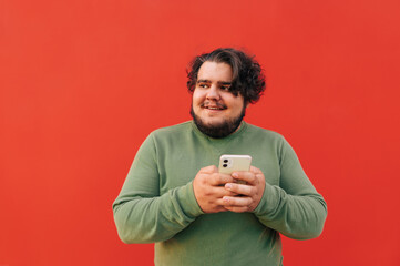 Young corpulent bearded hispanic guy is looking aside, holding and using his smartphone, feeling cheerful, standing on a red background.
