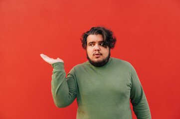 Fototapeta premium Funny and amusing young hispanic man is displaying the copy space, a red background, with his hand, advertising a product, trying to interest the viewer.
