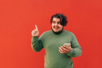 Attractive fat guy with a confused facial expression is pointing his finger up and holding a mobile...