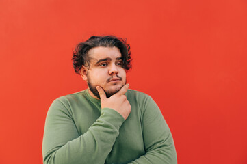 Closeup portrait of a thoughtful handsome bearded hispanic guy, touching his chin with his hand,...