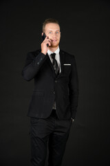 Portrait of young smart confident and successful businessman in black suit using smartphone isolated on black background