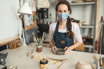 Caucasian woman making ceramic soup plate while wearing protective face mask for coronavirus prevention - Female person at work inside her creative pottery studio - Focus on face