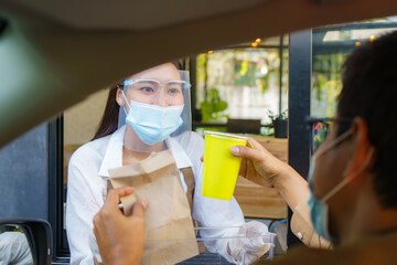 Asian man in protective mask taking food bag and coffee with woman waitress wearing face mask and face shield at drive thru during coronavirus outbreak..