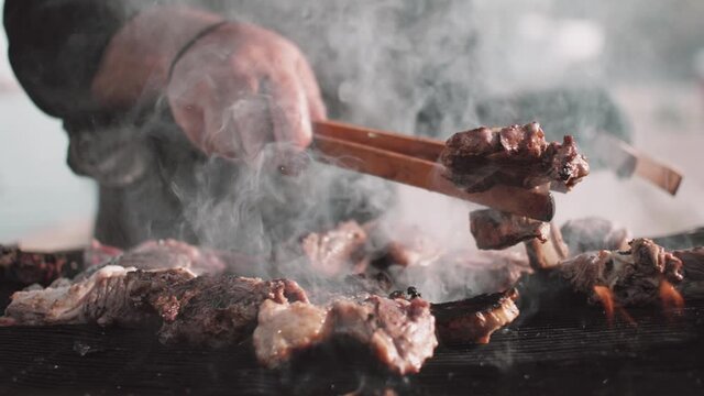 Chef uses a forceps to turn over smoky pork ribs cocking on barbecue. Process of cocking delicious meat outdoors on backyard