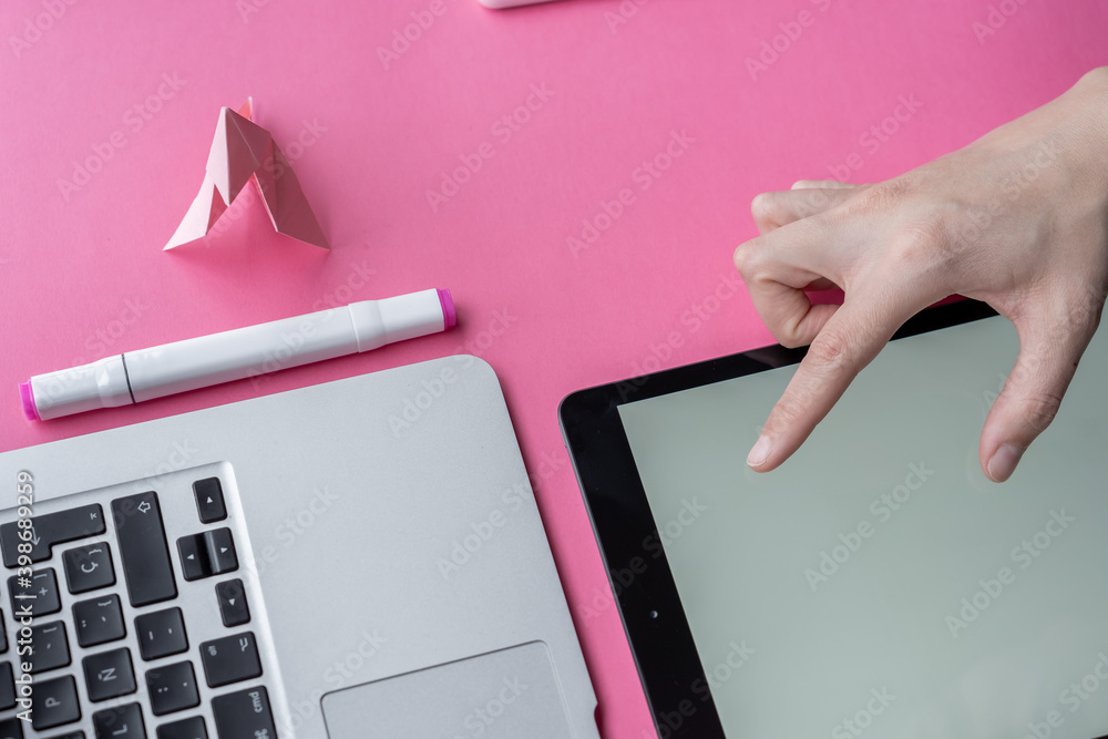 Wall mural Hand on a tablet with markers, notebook, laptop, swan origami, and smartphone on pink background