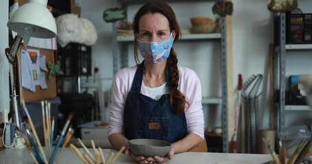 Caucasian woman making ceramic soup plate while wearing protective face mask for coronavirus prevention - Female person at work inside her creative pottery studio - Powered by Adobe