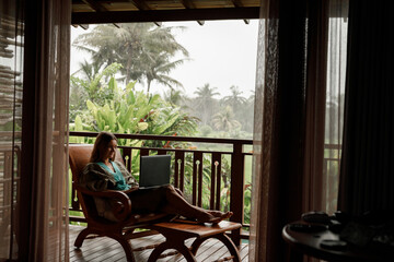 Freelance girl wering bathrobe  sitting on the balcony working, typing on laptop during raining...