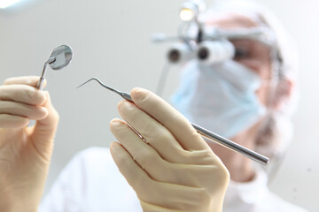 A doctor wearing binocular glasses holds a dental mirror and tweezers in his hand. The face is out of focus. Prevention of caries and gum diseases. Bottom view.
