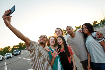 A group of six young commonly dressed friends having a good time together hugging each other and making a group selfie outside on a parking site with cars on a background