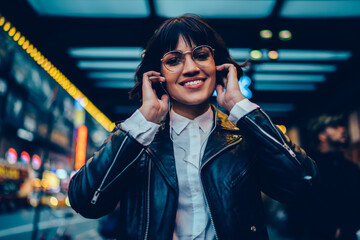 Half length portrait of cheerful female millennial in electronic spectacles smiling at camera during time for listening audio book and walk in metropolitan downtown, generation z in earphones