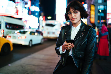 Portrait of millennial hipster girl in trendy wear holding smartphone standing on metropolitan...
