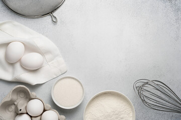 Top view of baking ingredients for cake. Wheat flour, sugar, whisk, sieve and white eggs on grey background. Copy space. 