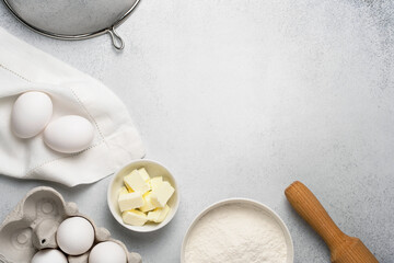 Top view of baking ingredients for dough. Wheat flour, butter, rolling pin, sieve and white eggs on grey background. Copy space. 
