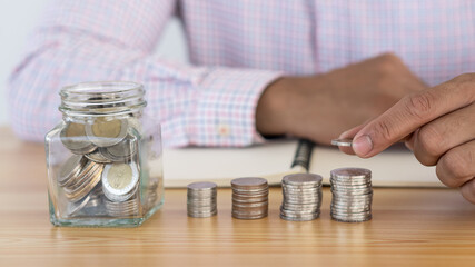 The piles of coins on the table and the glass jar with the hand held the coins save money in the future.