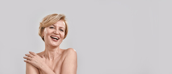 Portrait of beautiful middle aged woman laughing at camera, touching her skin, posing isolated against grey background. Beauty, skincare concept