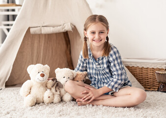 Little girl sitting with crossed legs near teddy bears on room floor with wigwam