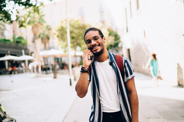 Cheerful dark skinned male tourist in casual wear making mobile phone call during journey using...