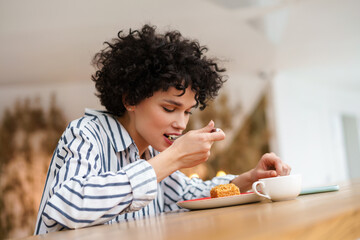 Beautiful pleased curly woman eating cake while drinking coffee in cafe