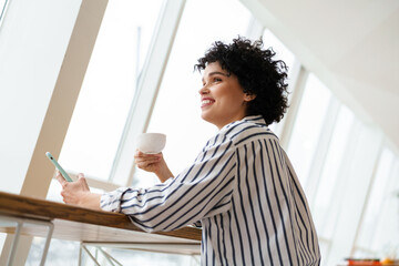 Fototapeta premium Beautiful smiling curly woman using mobile phone while drinking coffee