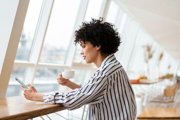 Beautiful focused curly woman using mobile phone while drinking coffee