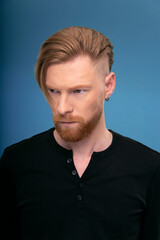 Fototapeta premium Handsome redhaired bearded man, wearing black shirt, posing frontal in studio, looking seriously, over blue background.